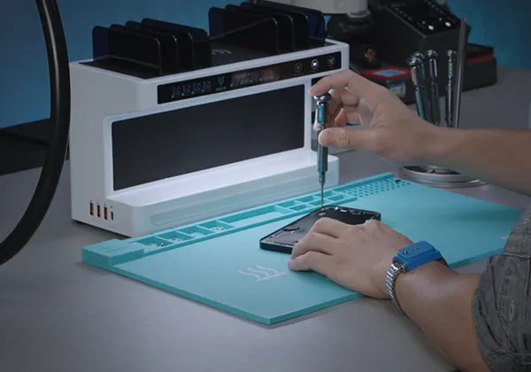 Person working on a phone with a tool in front of a white electronic device on a blue mat.