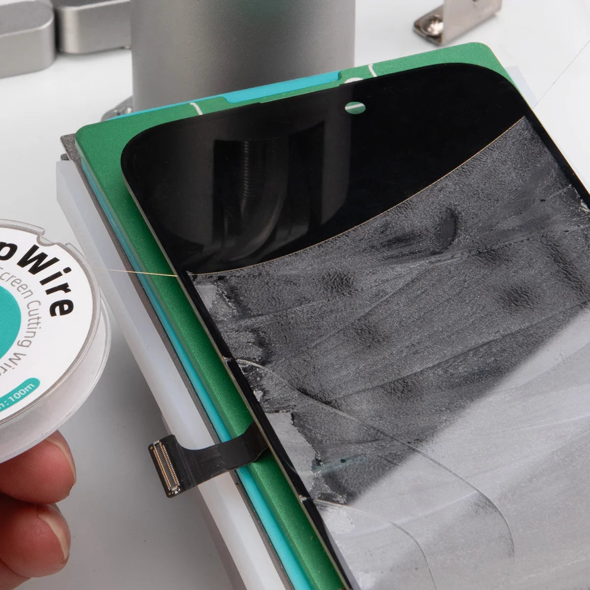 A close-up of a hand holding a spool of clear conductive wire next to a smartphone repair setup with a black screen protector and a green-and-white mounting board.