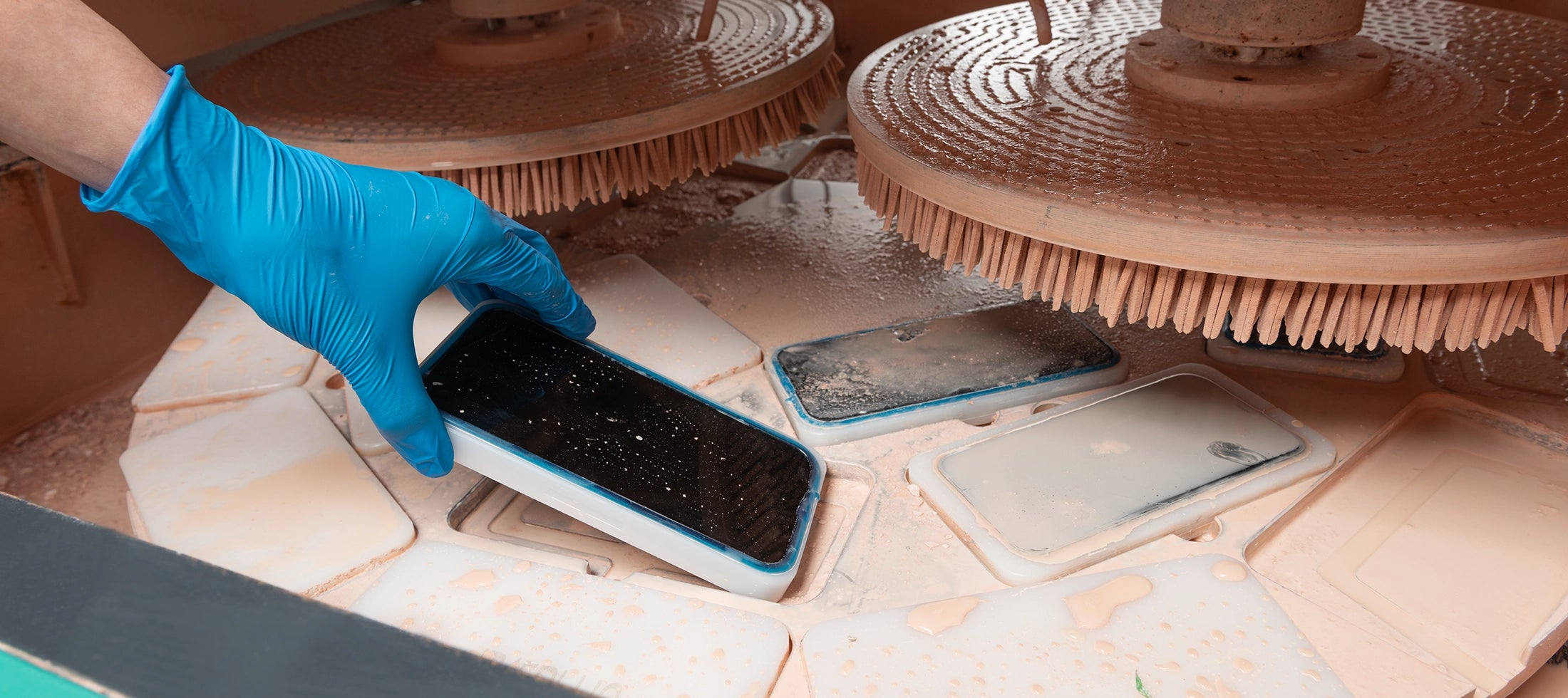 Person cleaning a smartphone with a brush and black substance on a workbench.